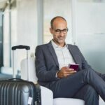Mature_businessman_using_mobile_phone_at_the_airport_in_the_waiting_room._Business_man_typing_on_smartphone_in_lounge_area._Portrait_of_latin_man_sitting_and_holding_passport_with_luggage.