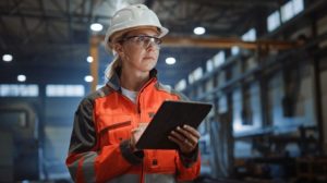 Worker_Wearing_Safety_Uniform_and_Hard_Hat_Uses_Tablet_Computer._Serious_Successful_Female_Industrial_Specialist_Walking_in_a_Metal_Manufacture_Warehouse.