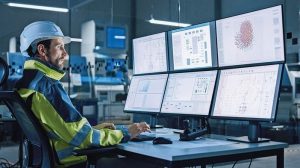 Industry_4.0_Modern_Factory:_Facility_Operator_Controls_Workshop_Production_Line,_Uses_Computer_with_Screens_Showing_Complex_UI_of_Machine_Operation_Processes,_Controllers,_Machinery_Blueprints