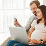 Portrait_of_a_happy_young_pregnant_couple_shopping_online_while_sitting_on_a_couch_with_credit_card_and_laptop_computer