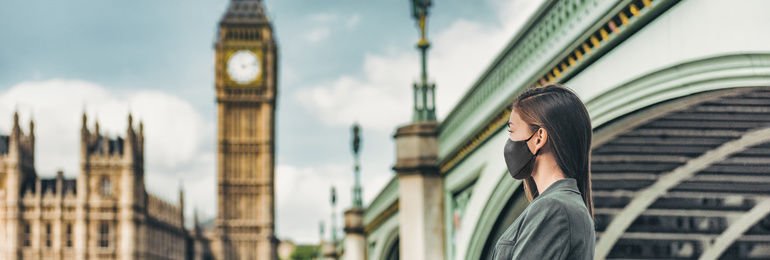 COVID-19_mask_wearing_business_people_walking_in_London_city._Asian_woman_using_face_cover_for_public_outdoor_spaces_in_urban_landscape_banner._Tourist_at_view_of_Big_Ben,_UK.