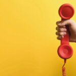 Closeup_view_of_woman_holding_red_corded_telephone_handset_on_yellow_background,_space_for_text._Hotline_concept